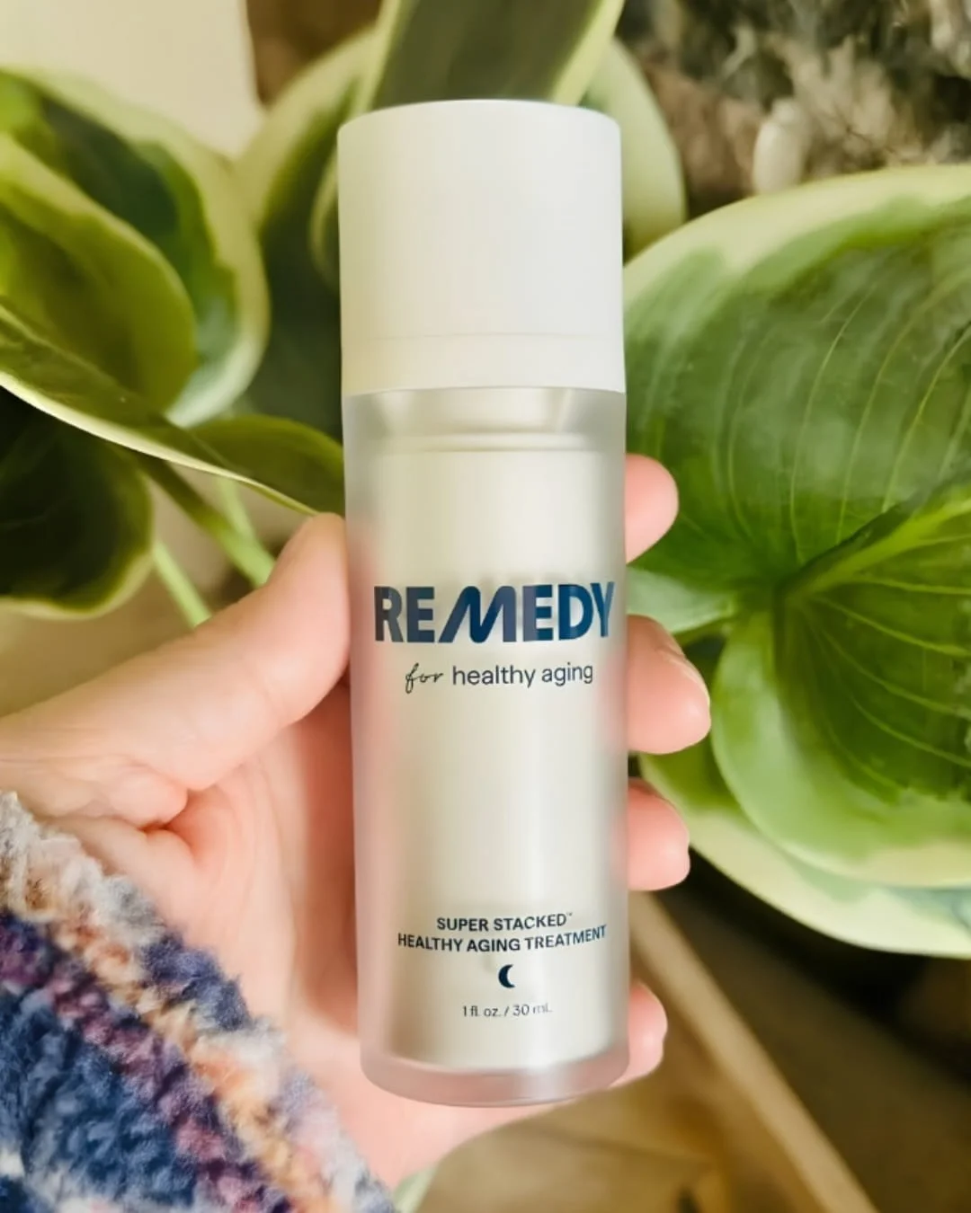 Hand holding a Remedy for healthy aging Super Stacked Healthy Aging Treatment bottle with a white cap and frosted body, shown in front of lush green leaves