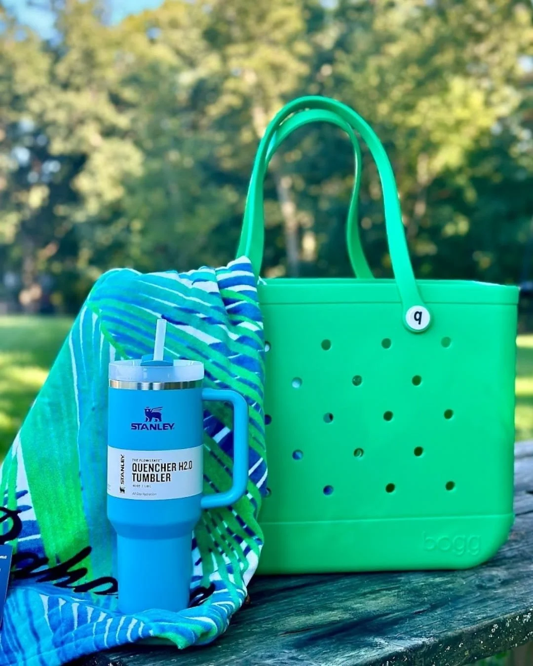Stanley Quencher H2.0 tumbler with handle and straw, vibrant green Bogg Bag with perforated design, and a colorful towel on a wooden surface against a natural outdoor background.