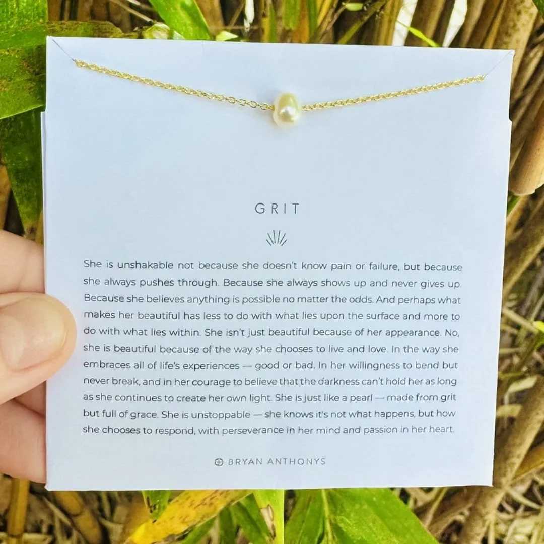 Bryan Anthonys Grit necklace featuring a gold chain with a single baroque pearl, displayed on a white card with an inspirational message about resilience and inner beauty, held against a natural green background.