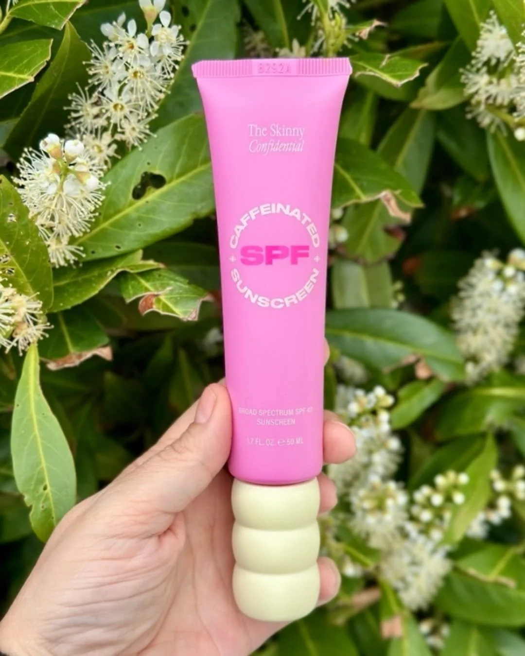 Bright pink tube of The Skinny Confidential Caffeinated Sunscreen SPF 40 with a cream-colored ridged cap, held upright in a hand against a lush green background with white flowers.