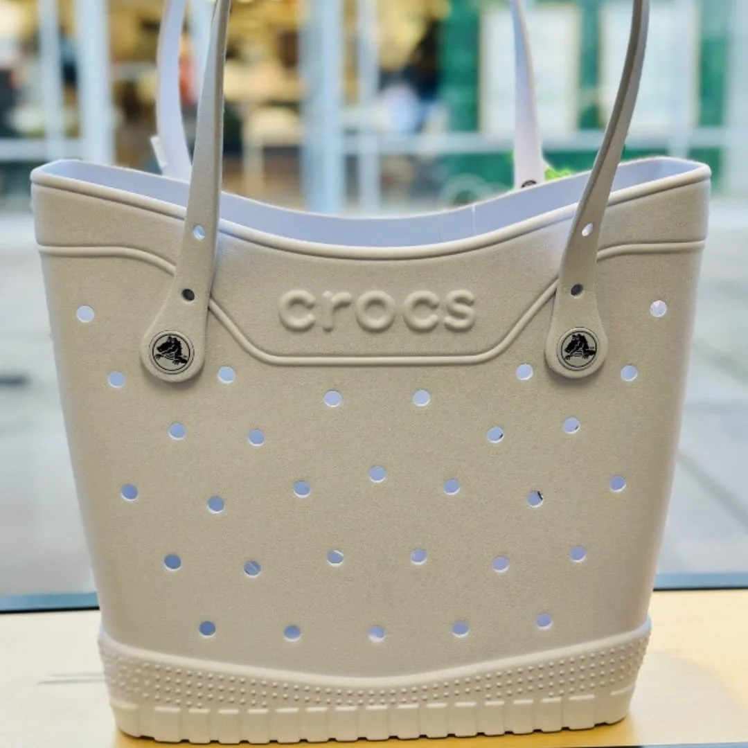 White Crocs-branded tote bag featuring ventilation holes, embossed logo, and textured base resembling a Crocs shoe, displayed on a shelf in a brightly lit retail store.