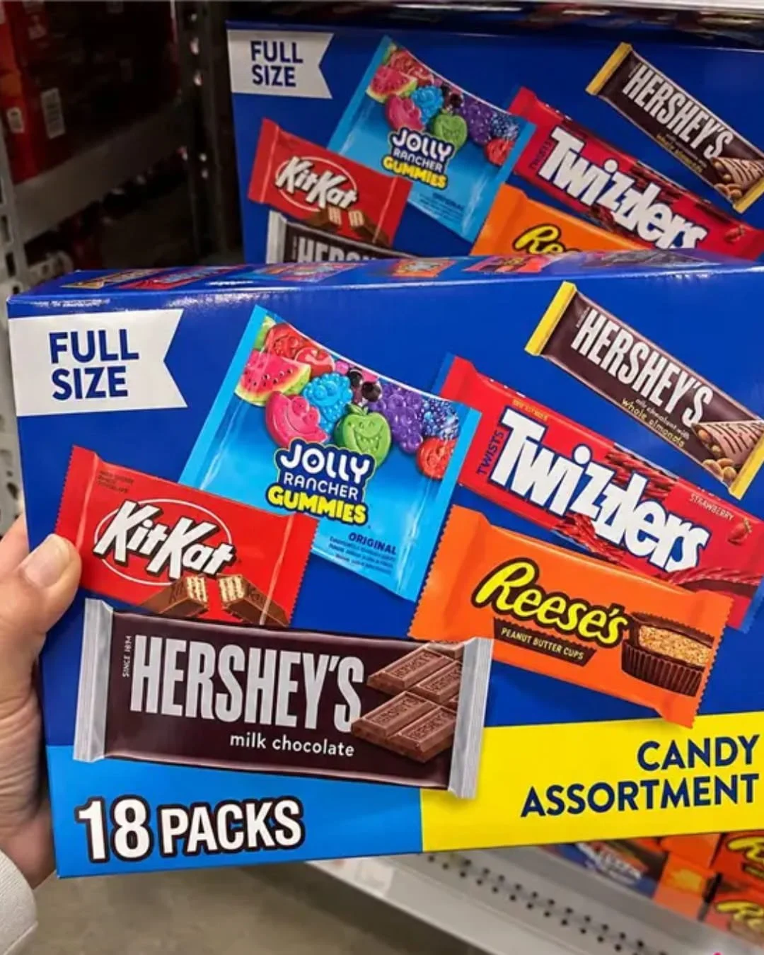 Hand holding a Hershey full size candy assortment 18-pack box featuring KitKat, Reese’s, Twizzlers, Jolly Rancher Gummies, and Hershey’s bars