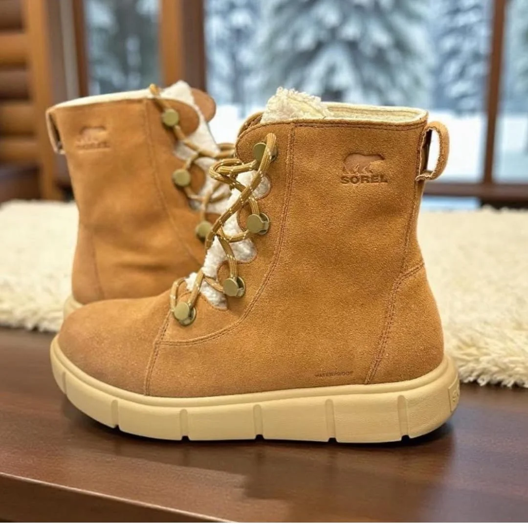 Brown Sorel winter boots with waterproof suede upper, fluffy cream lining, and chunky beige soles, placed on a wooden surface with a white rug, set against a snowy outdoor background.