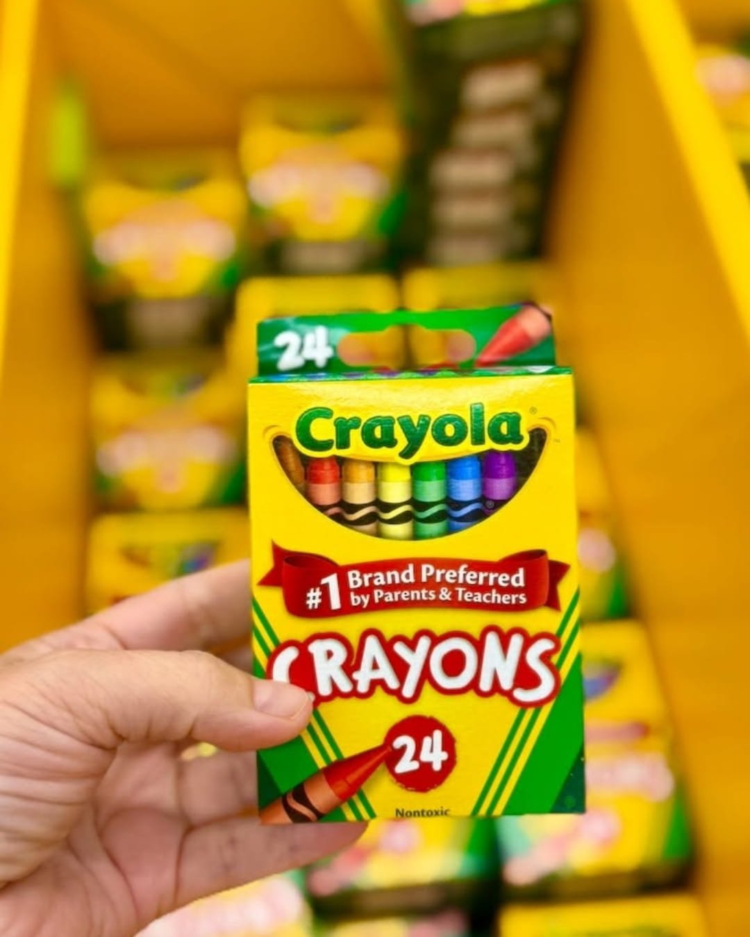Hand holding a 24-count box of Crayola crayons with colorful crayon tips visible through the front window in a store aisle
