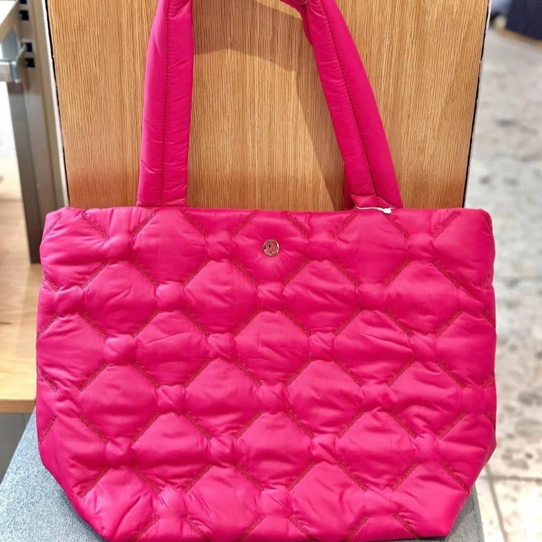 Fuchsia pink Lululemon quilted tote bag with diamond stitching, padded handles, and a metallic logo, displayed on a gray surface against a wooden background.