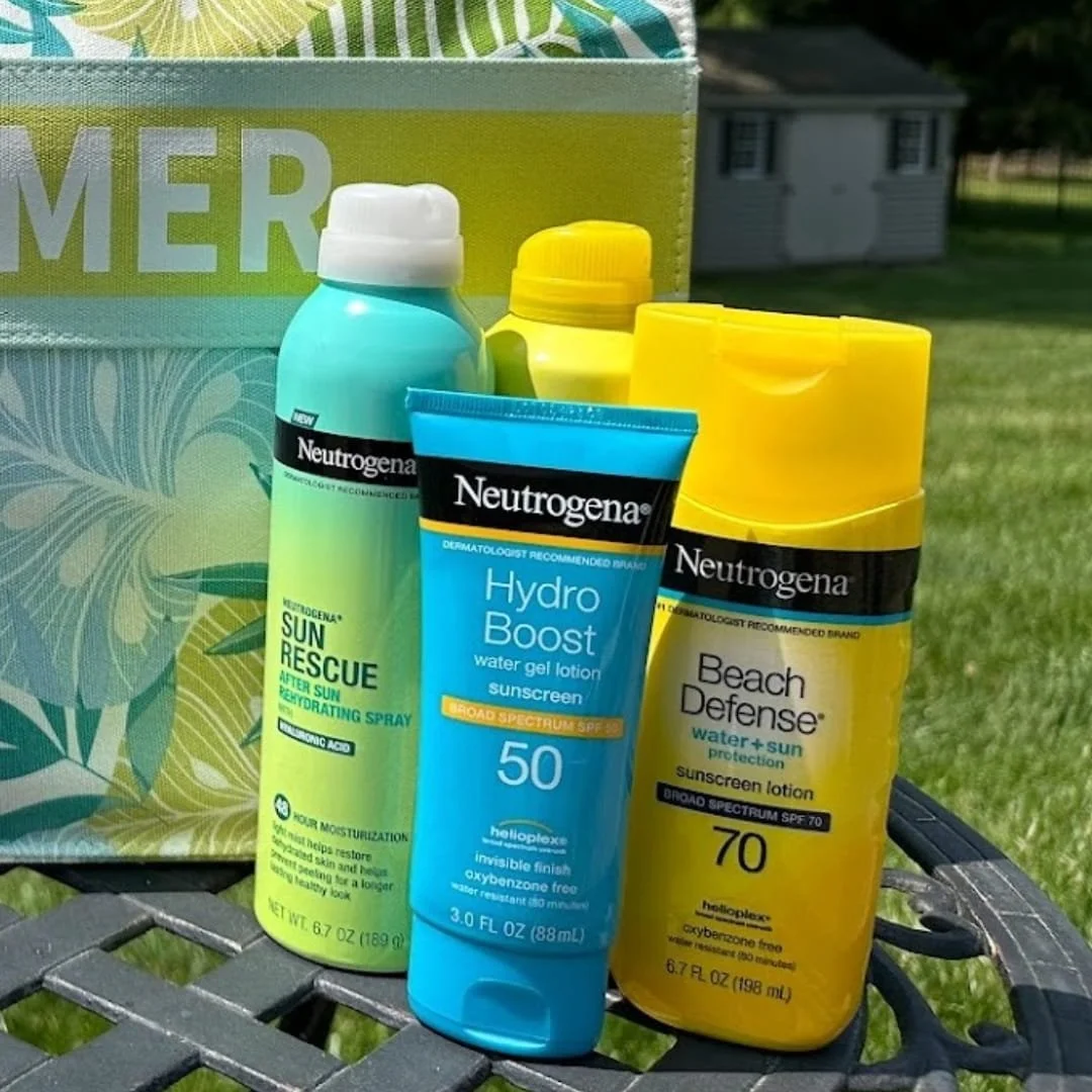 Neutrogena sun care products including Sun Rescue After Sun Spray, Hydro Boost SPF 50 water gel lotion, and Beach Defense SPF 70 sunscreen lotion, displayed on a patio table with a tropical-themed bag in the background.
