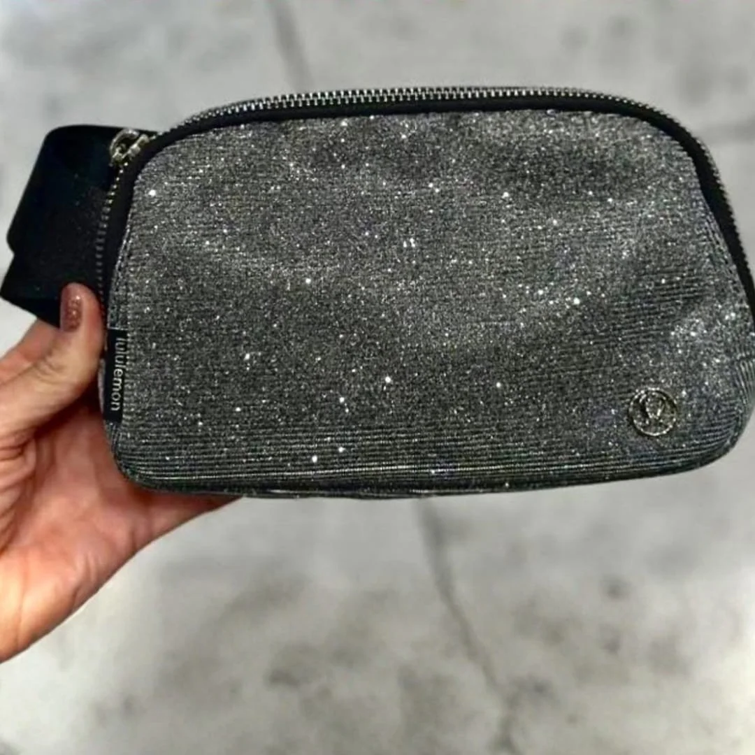 Lululemon glittery dark belt bag with a metallic logo, black zipper, and strap, held by a hand with manicured nails against a light textured background.