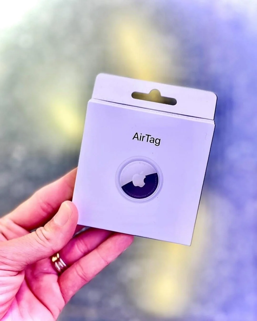 A hand holding a white Apple AirTag product box, featuring the AirTag name and a circular graphic of the device with the Apple logo, against a softly blurred background.