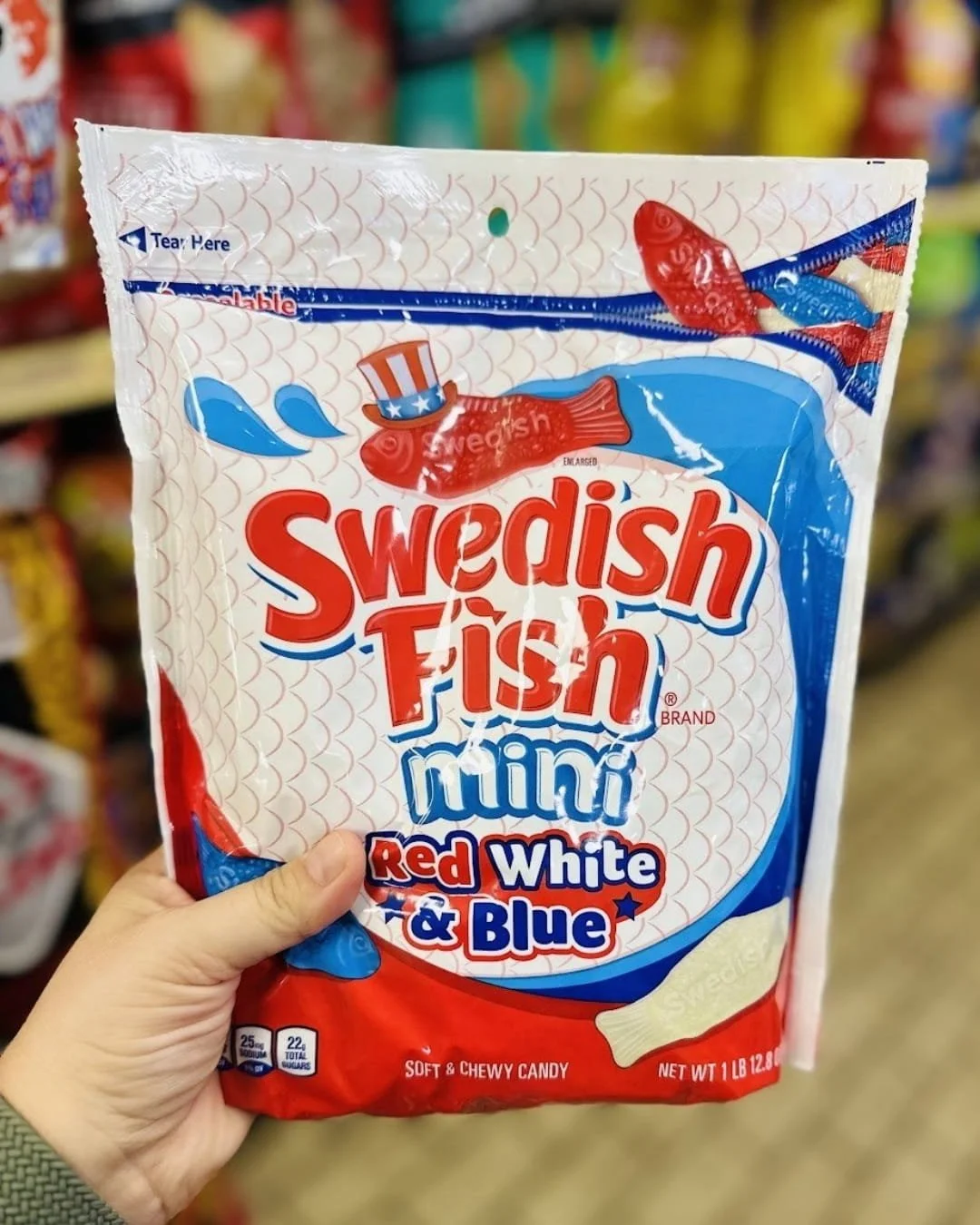 Bag of Swedish Fish Mini Red White & Blue gummy candy featuring patriotic-themed packaging with a red fish wearing an Uncle Sam hat, held in a hand against a blurred grocery store shelf background.