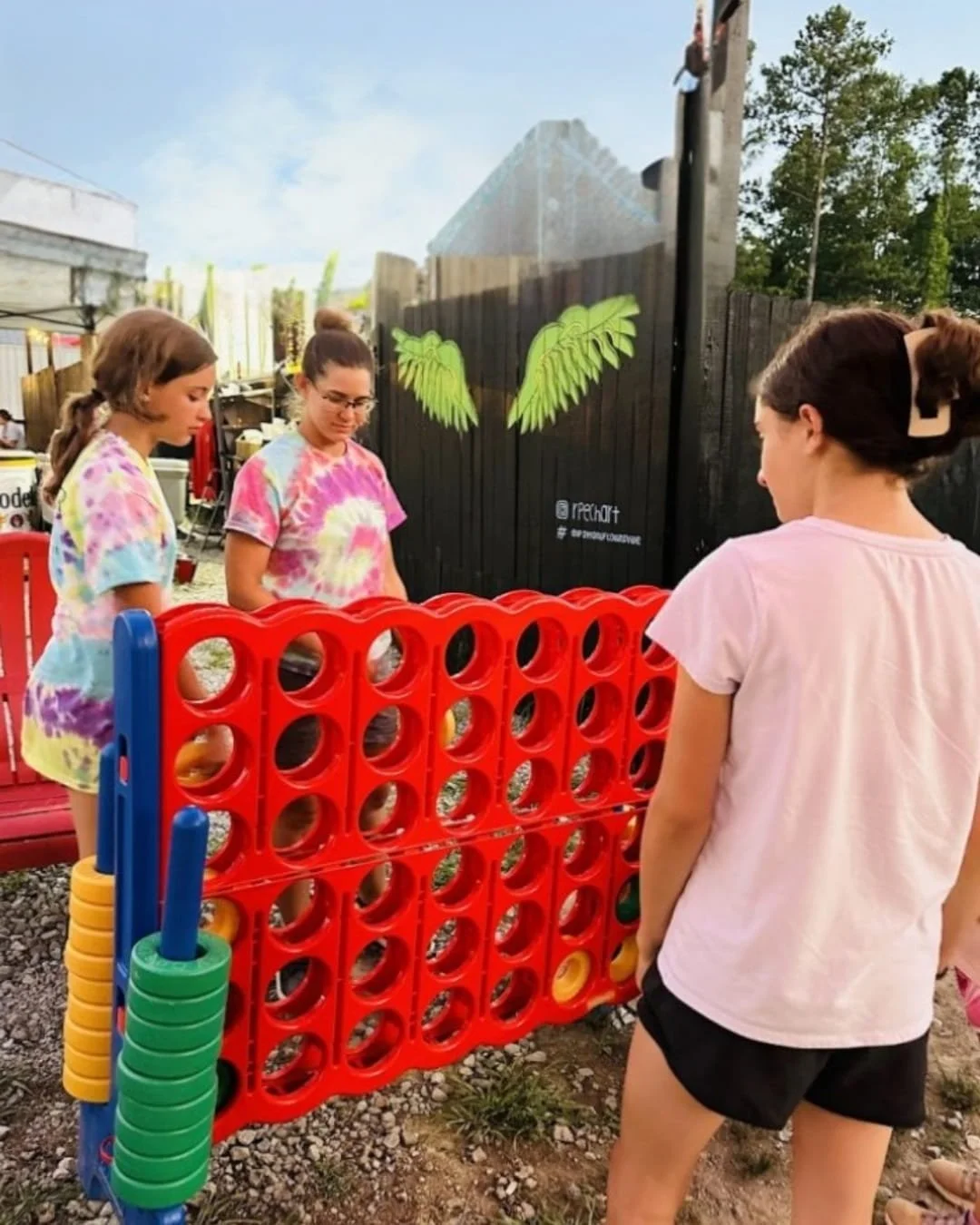 Three young women playing a giant Connect Four game outdoors, with a red game board, blue legs, and yellow and green rings, set against a gravel ground, a mural fence with green wings, and a backdrop of trees and sky.