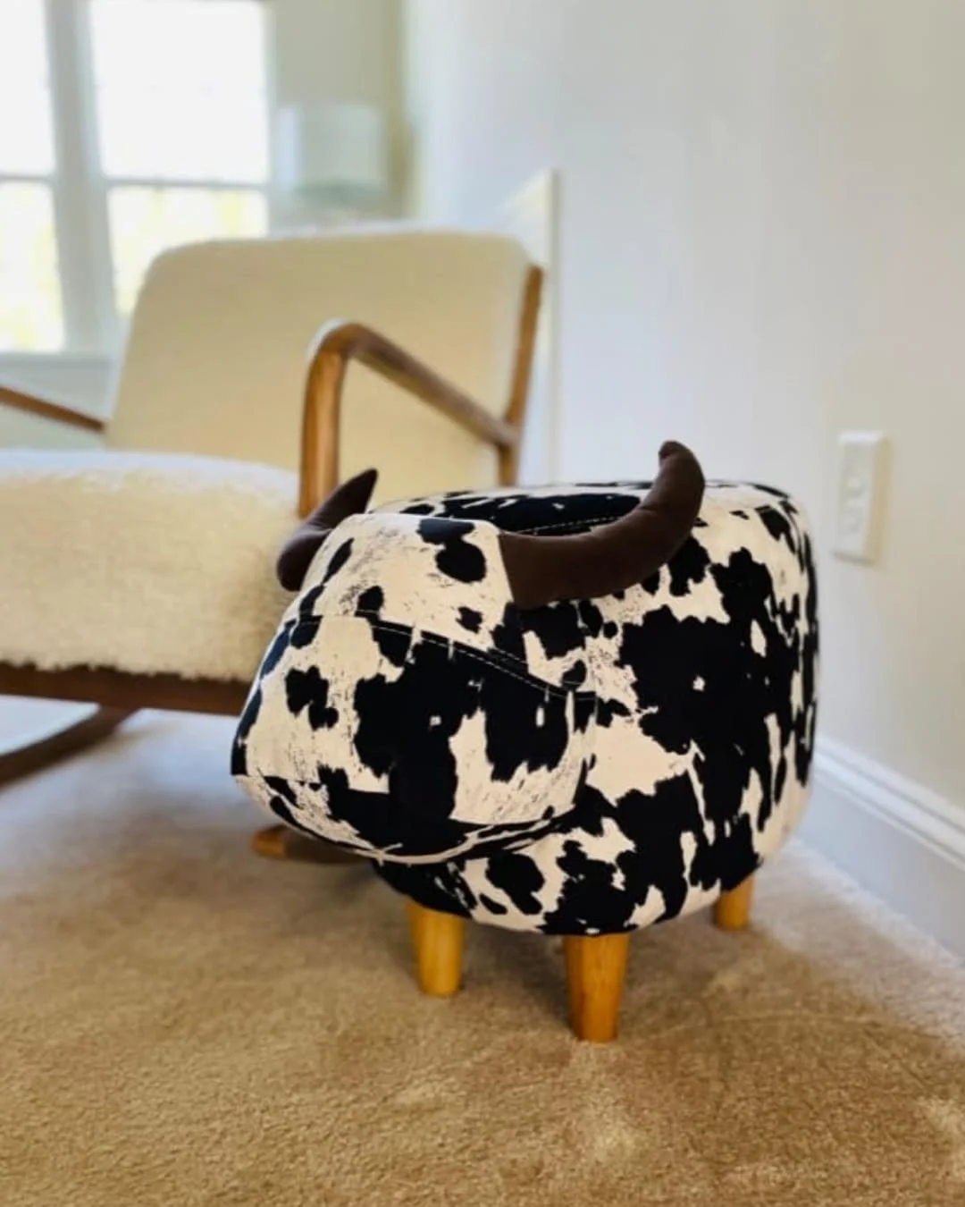 Cow-print ottoman with plush horns and wooden legs in a bright, cozy room, paired with a sherpa-upholstered rocking chair on a beige carpet.