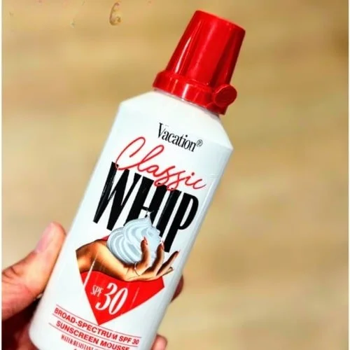 "White can of Vacation Classic WHIP Sunscreen Mousse with a red cap, SPF 30 label, and a retro-style graphic of a hand holding whipped mousse, held against a blurred beige background.