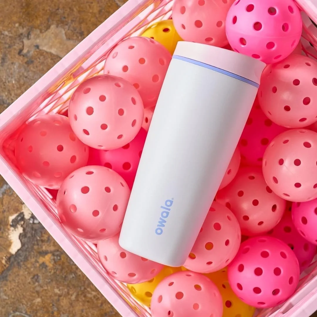 White Owala tumbler with a pink lid and blue logo, resting diagonally on a pile of pink and yellow pickleballs inside a pink crate, set against a neutral-toned background.