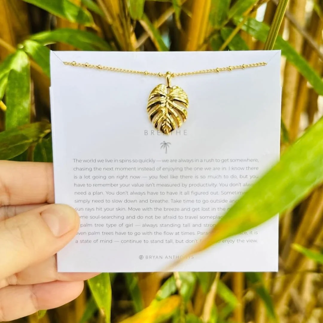 Gold leaf-pendant necklace from Bryan Anthony's draped over a white card with an inspirational message about mindfulness and resilience, held against a blurred green foliage background.