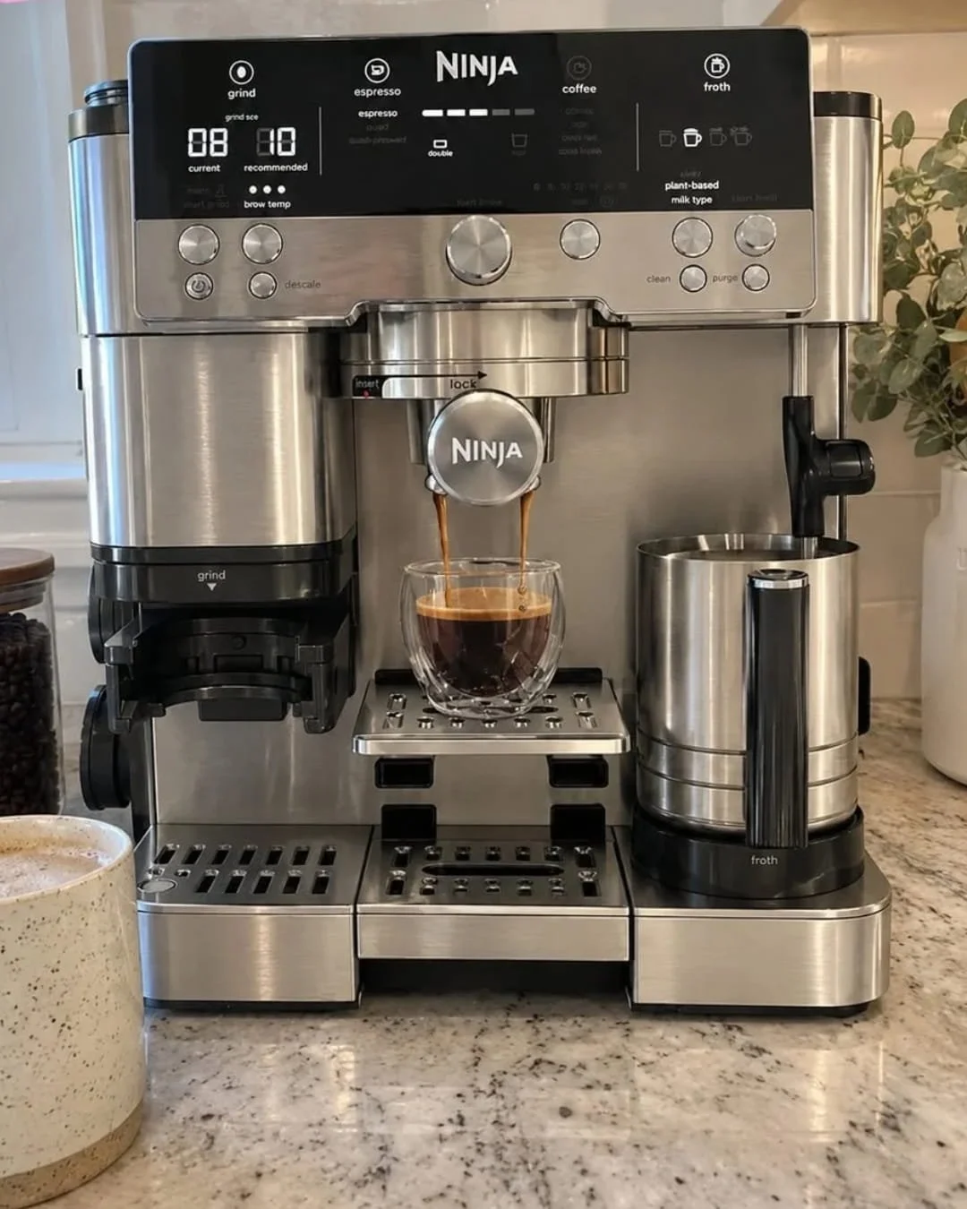 Ninja espresso machine brewing fresh espresso into a glass cup on a kitchen countertop with milk frother pitcher beside it