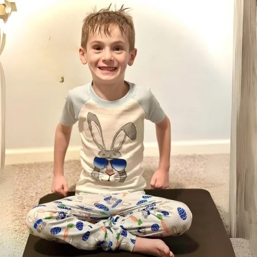 Smiling young boy sitting cross-legged on a dark mat, wearing Easter-themed pajamas with a bunny graphic and colorful egg and carrot patterns, against a light-colored wall.