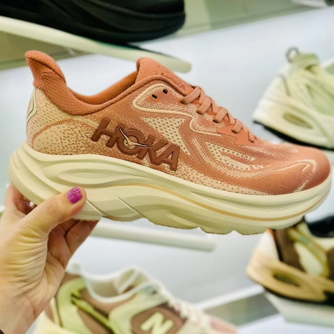 HOKA Clifton 10 running shoe in dusty peach with a thick cream cushioned sole, held in hand in a retail store