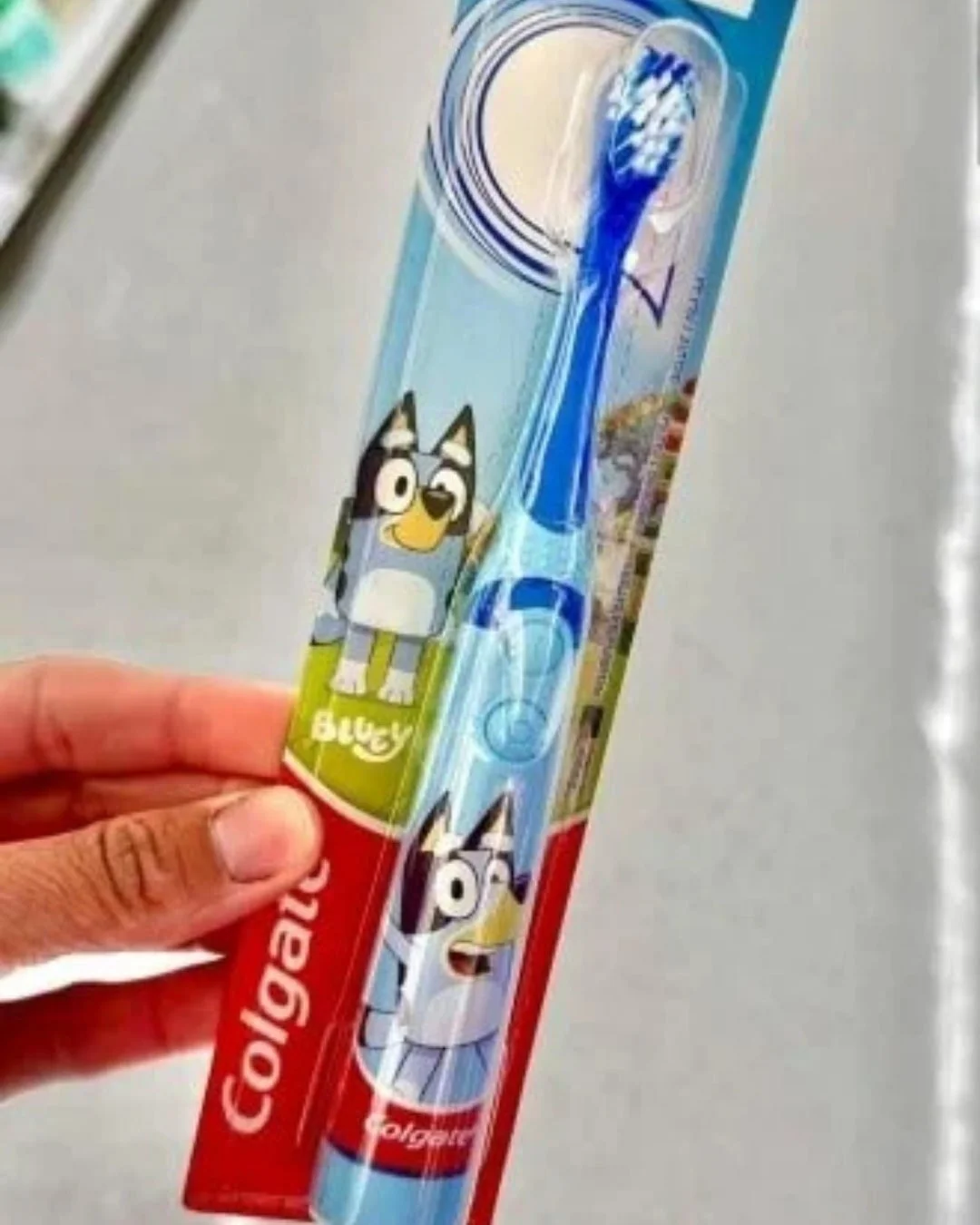 Colgate Bluey-themed children's toothbrush in blue and white, featuring Bluey illustrations on the handle and packaging, held by a hand against a blurred retail background.