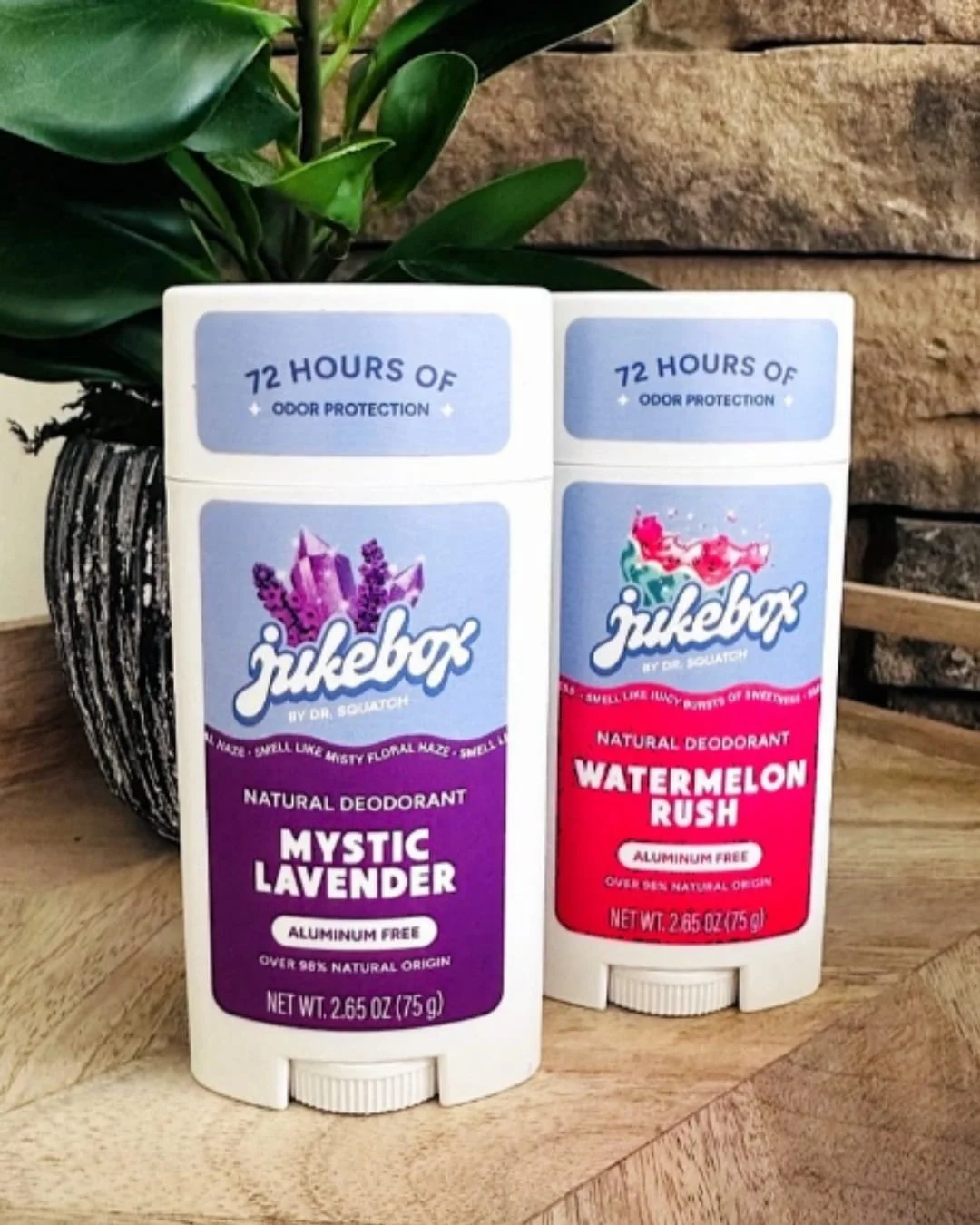 Jukebox by Dr. Squatch Natural Deodorants in Mystic Lavender and Watermelon Rush scents, featuring vibrant purple and pink packaging, displayed on a wooden surface with a green potted plant and a stone wall in the background.