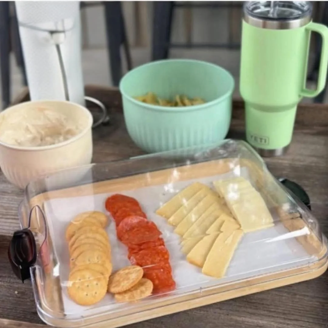 Casual charcuterie board with crackers, pepperoni, cheese slices, and dip in a clear-lidded tray, accompanied by a mint green YETI tumbler and a bowl of chips on a wooden table.