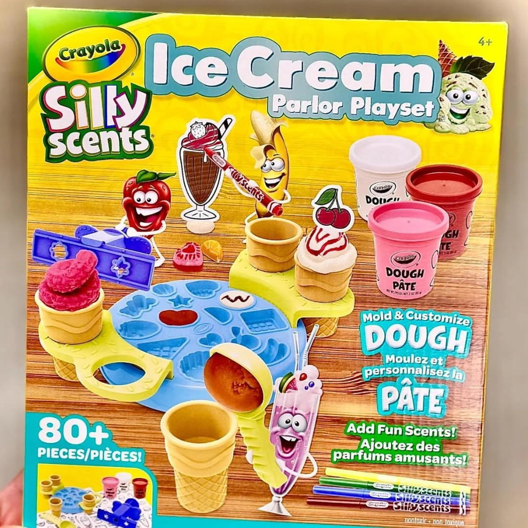 Crayola Silly Scents Ice Cream Parlor Playset featuring 80+ pieces, including molding tools, scented dough, and ice cream cones, displayed on vibrant packaging with playful characters.