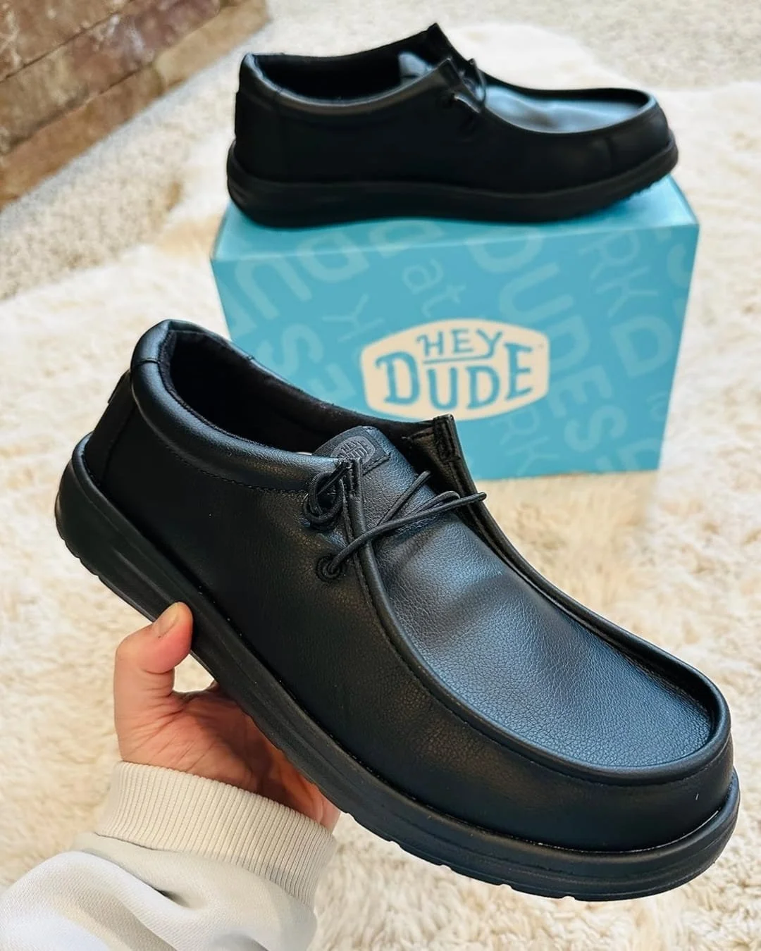 Hey Dude men's black loafers with moc-toe design, shown with one shoe held in hand and the other resting on a blue branded box, set on a cream shag rug.