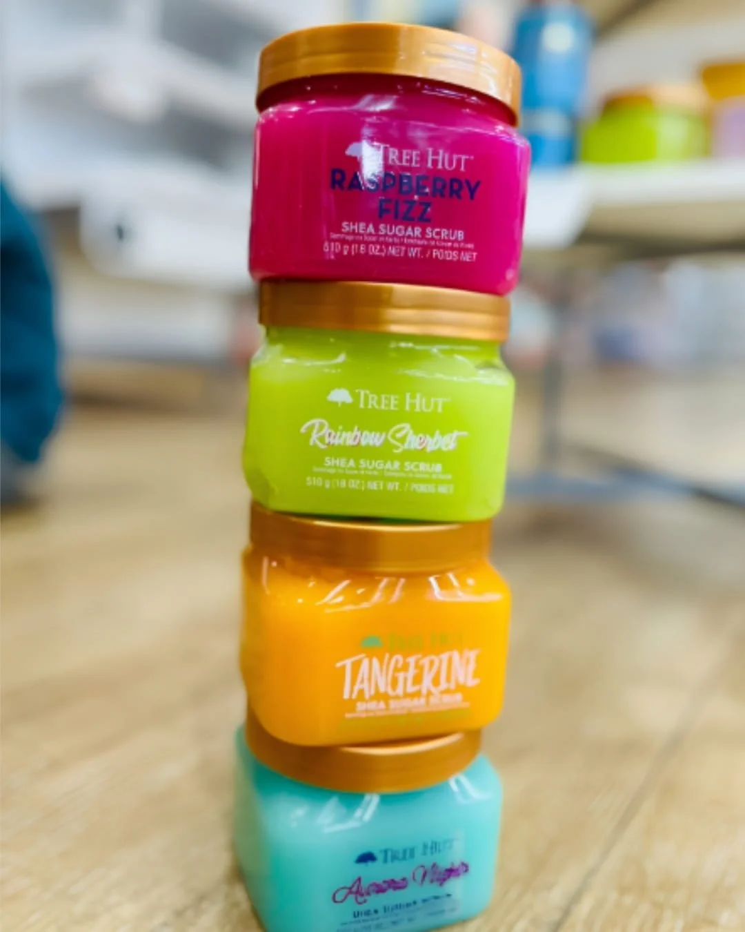 Stack of colorful Tree Hut Shea Sugar Scrub jars in multiple scents with gold lids on a wooden floor