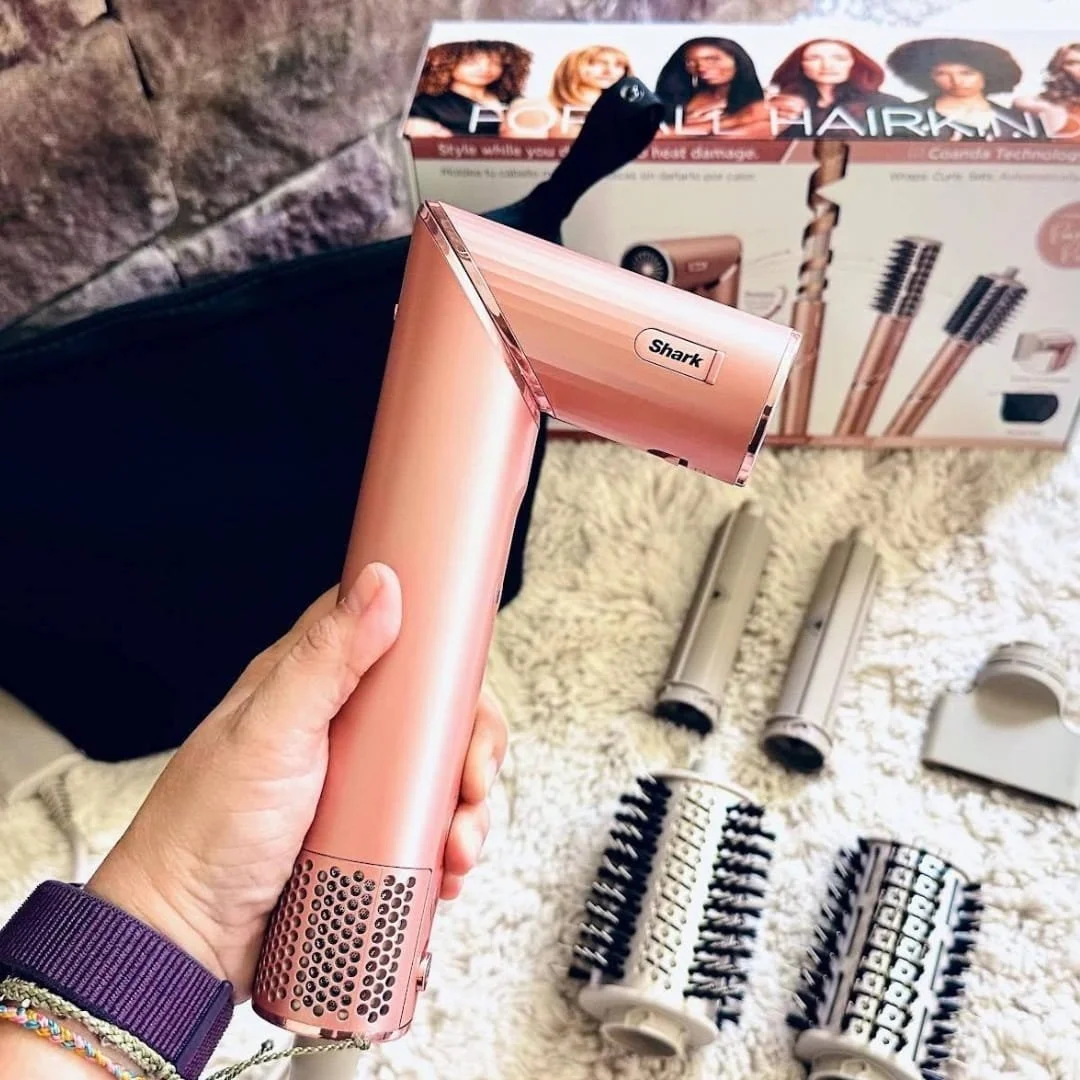 Rose gold Shark hair styling device with multiple attachments, including curling barrels and brushes, displayed on a fluffy white rug with product packaging in the background highlighting features for all hair types and heat damage protection.
