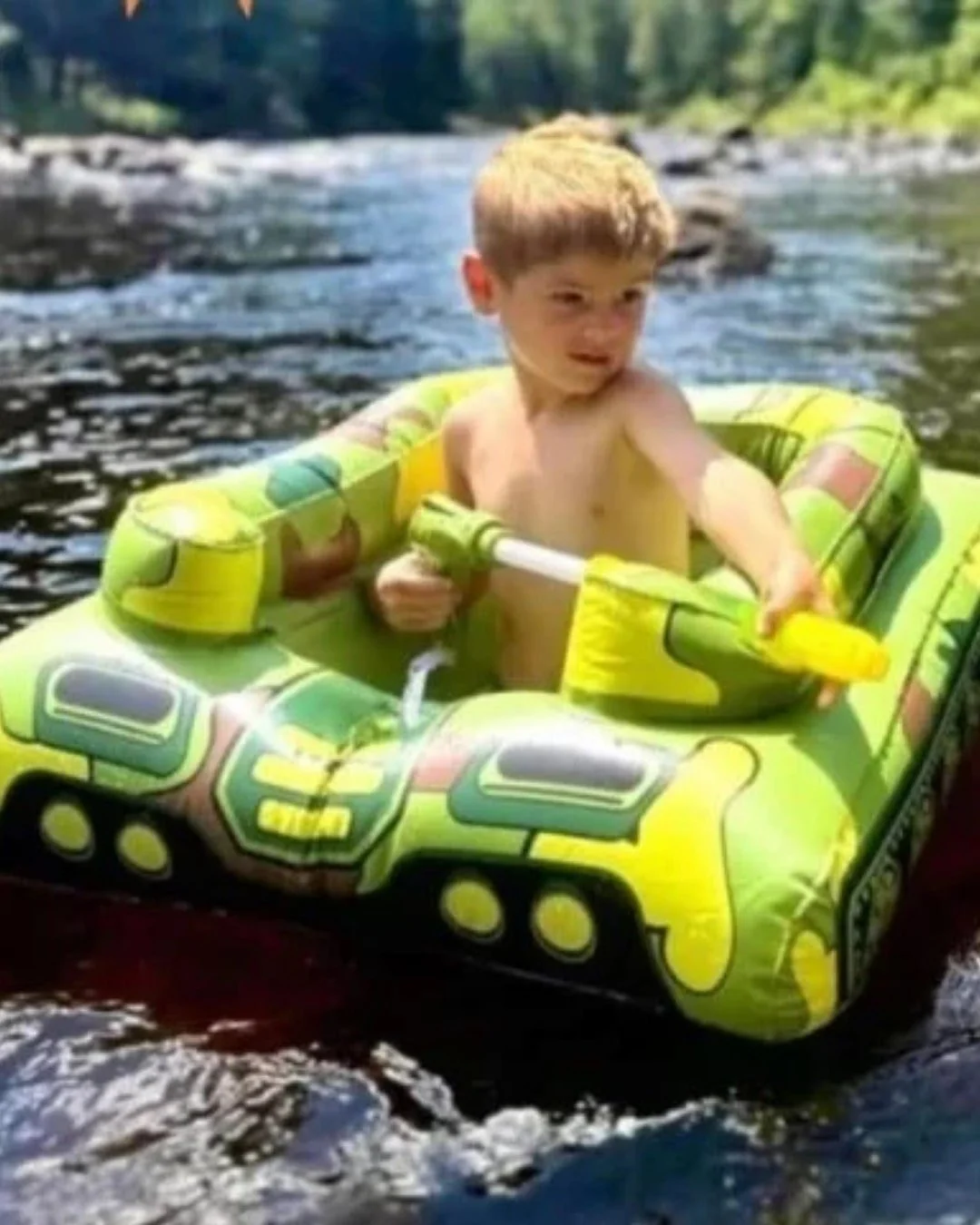 Young boy sitting in a green camouflage inflatable tank float in a body of water, holding a yellow and green water squirter, with a forested shoreline in the background.