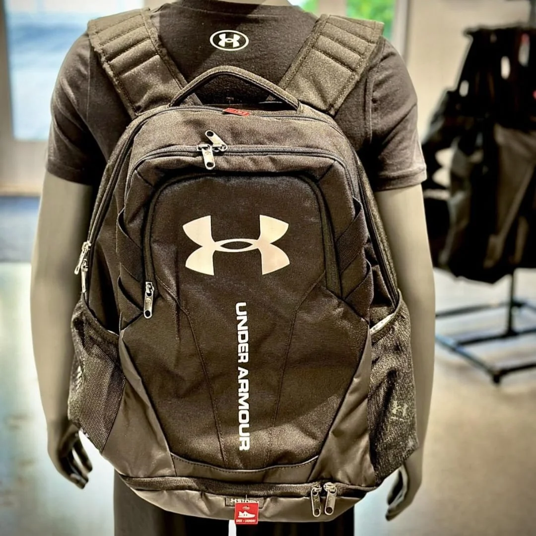 Black Under Armour Storm backpack with water-resistant features, displayed on a mannequin wearing a matching Under Armour t-shirt in a retail store setting.