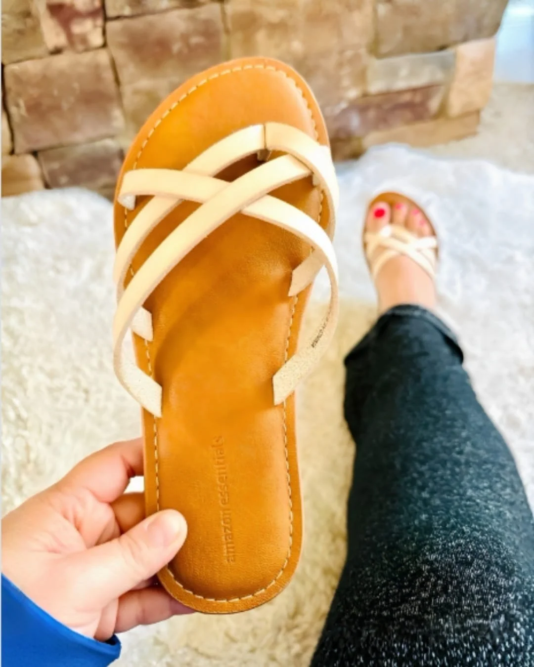 Hand holding a women’s slide flat sandal with cream crisscross straps and a tan footbed, with the matching sandal worn on foot in the background on a white textured rug