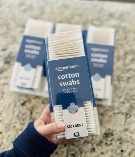 Hand holding a clear plastic package of Amazon Basics cotton swabs with a blue and white label displaying 500 COUNT,  Double Tipped, and 100% Cotton, with additional packages blurred in the background on a speckled countertop.