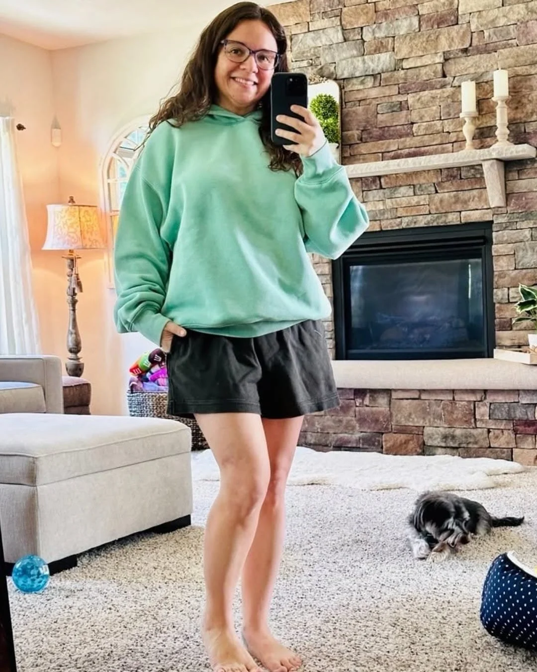 Nicole taking a mirror selfie while wearing an AE Everyday Luxe mint green oversized hoodie with dark shorts, standing barefoot in a cozy living room with a stone fireplace and dog in the background