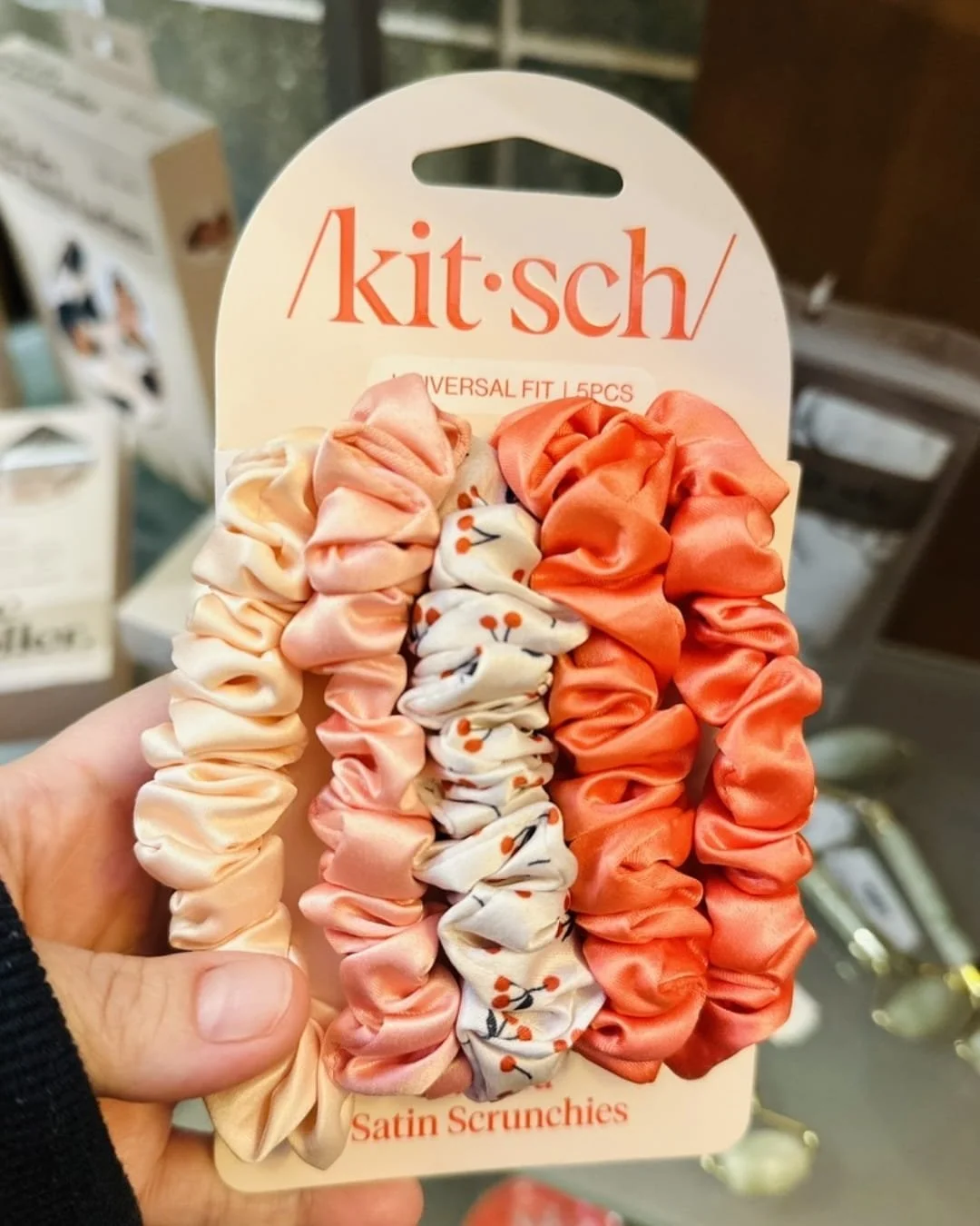 Kitsch Satin Scrunchies 5-pack in warm tones including cream, peach, cherry print, coral, and burnt orange, displayed on white packaging held by a hand in a retail setting.