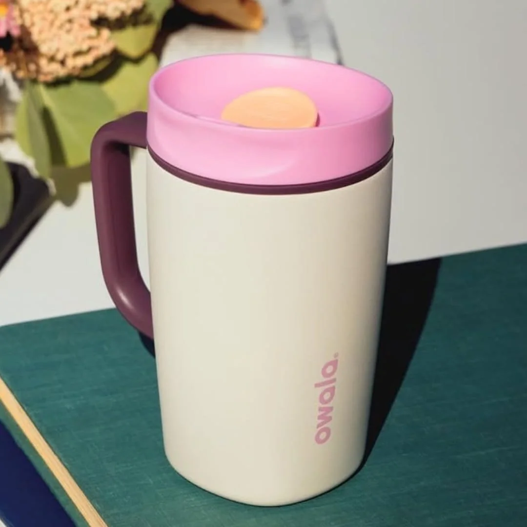 Owala travel mug with cream body, purple handle, and pink lid styled on a dark green book