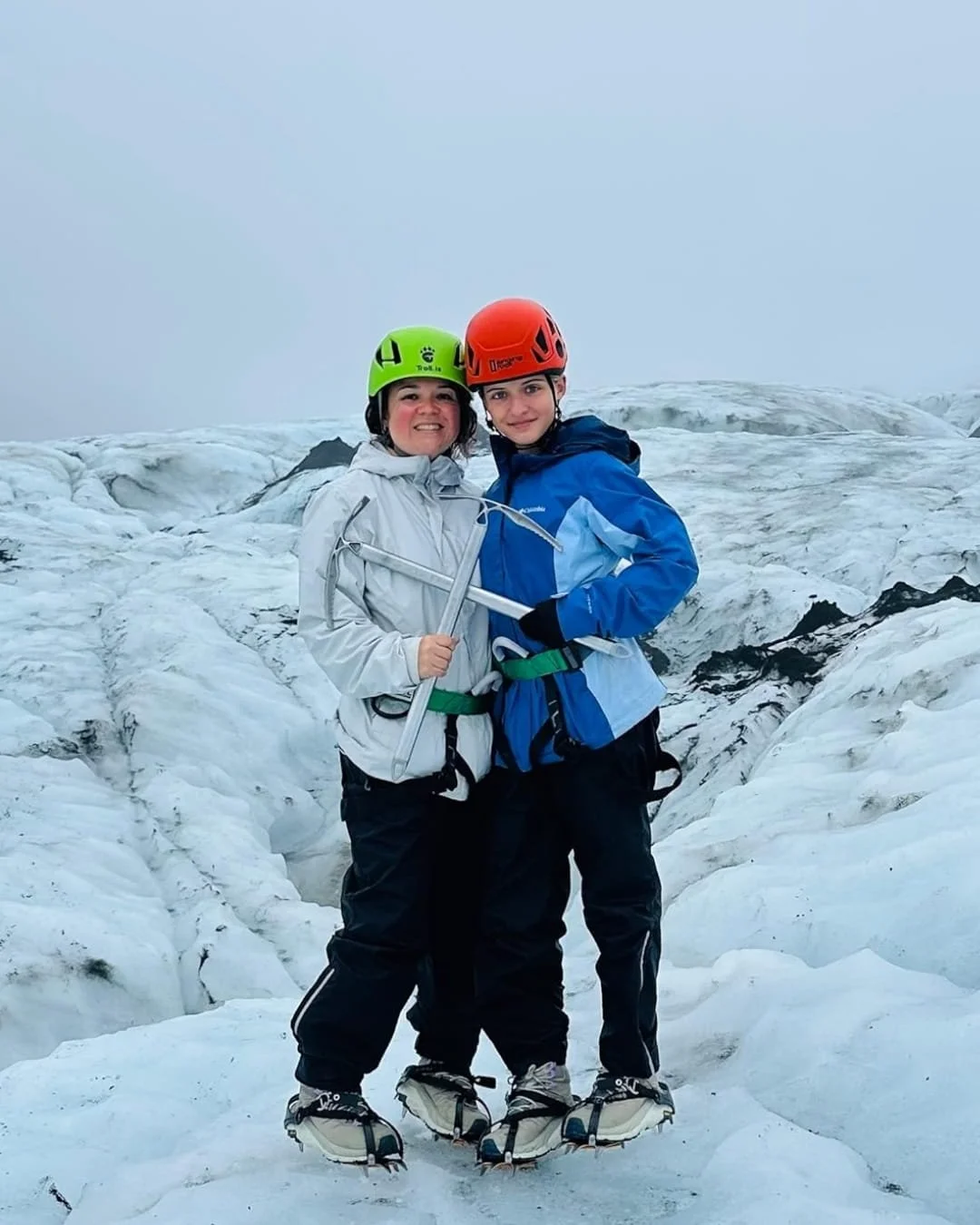 Two individuals on a glacier hike, wearing technical gear including a Columbia women’s blue jacket, helmets, harnesses, and crampons, standing on a rugged ice field with overcast skies in the background.