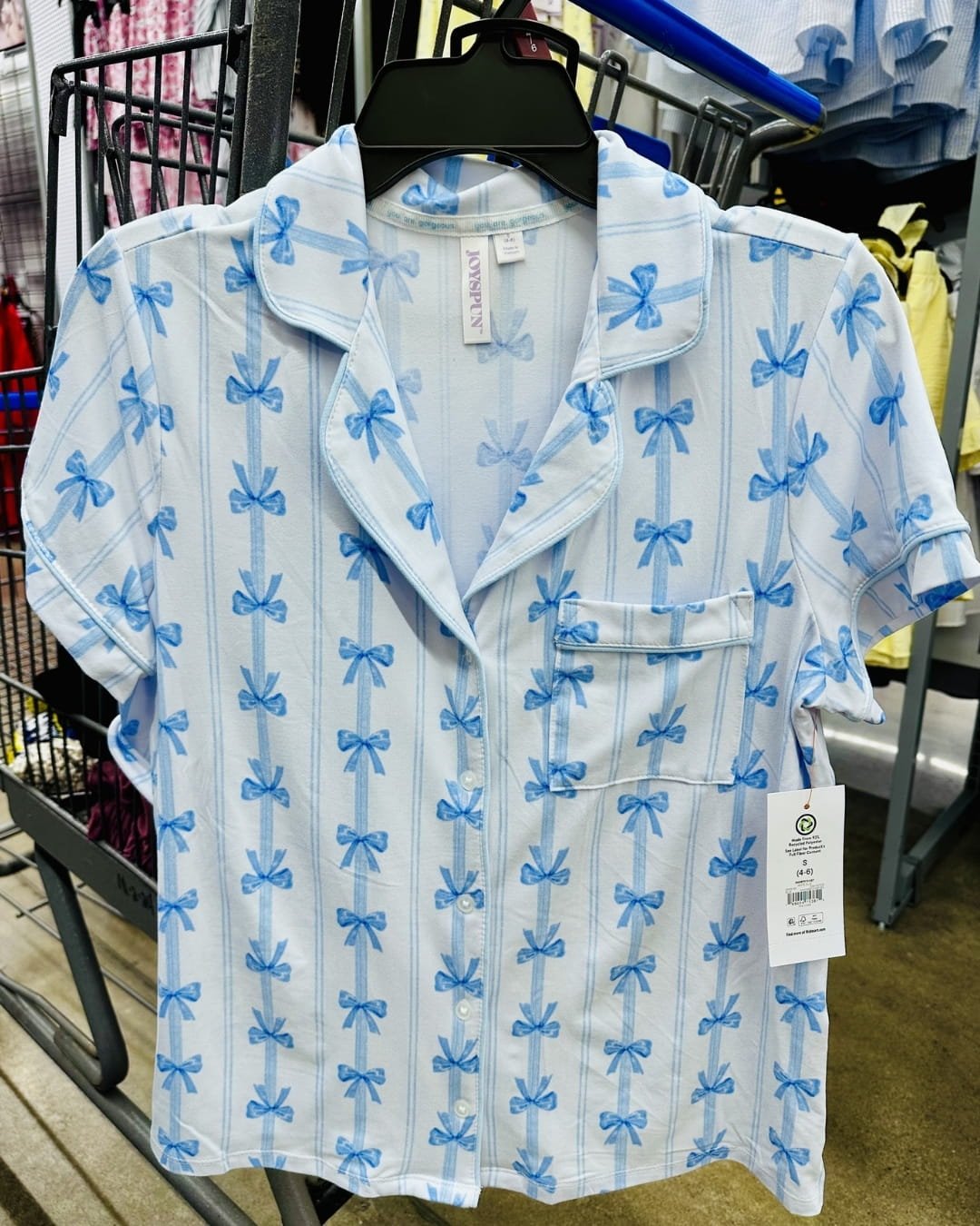 Joyspun short-sleeved pajama top with light blue bow and stripe pattern, hanging on a black hanger in a retail store, featuring a notched collar, chest pocket, and sustainable material tag.