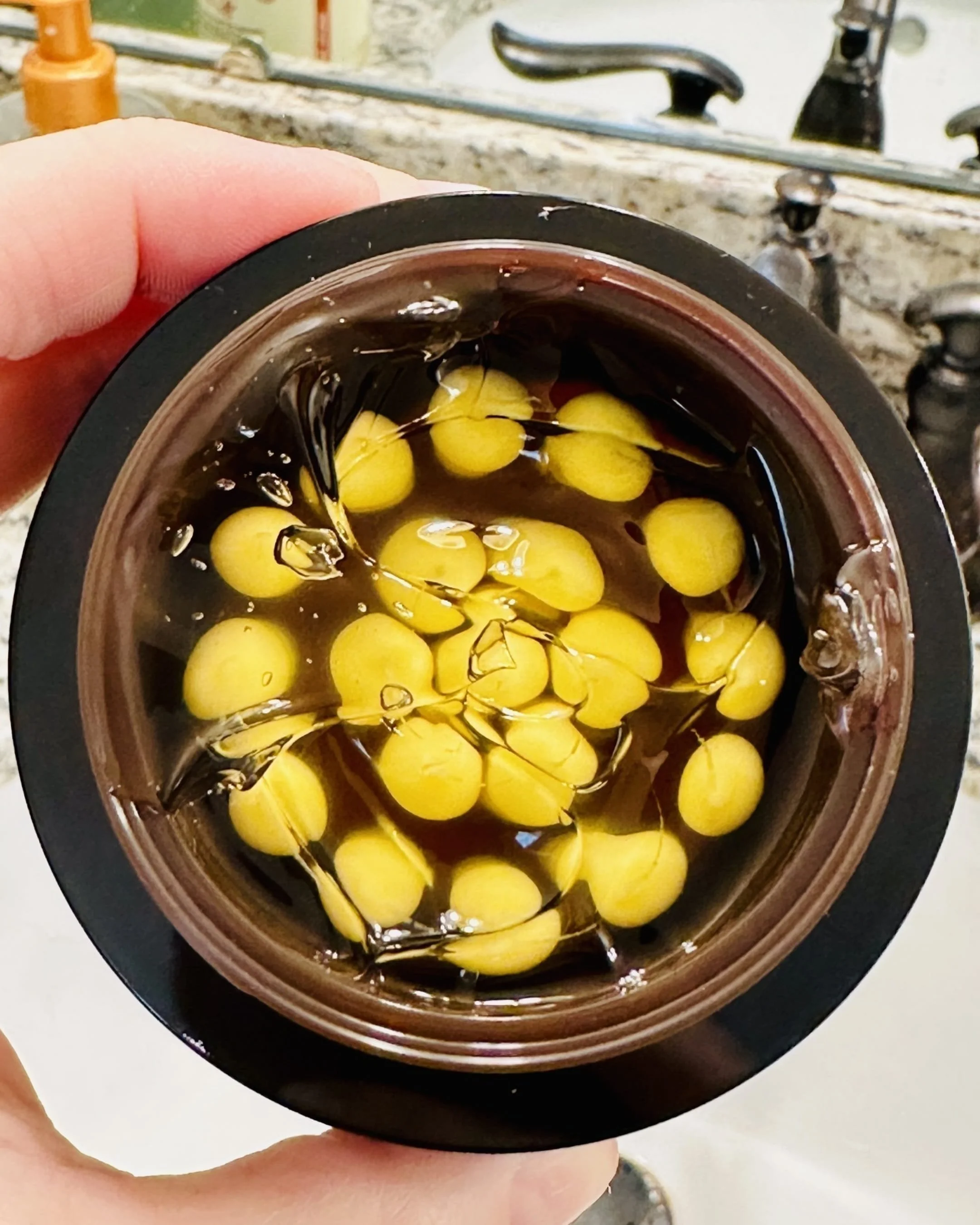 Open jar of Medicube Capsule Cream featuring amber-colored gel with suspended pale yellow capsules, held over a bathroom sink with a granite countertop and metallic faucet in the background.