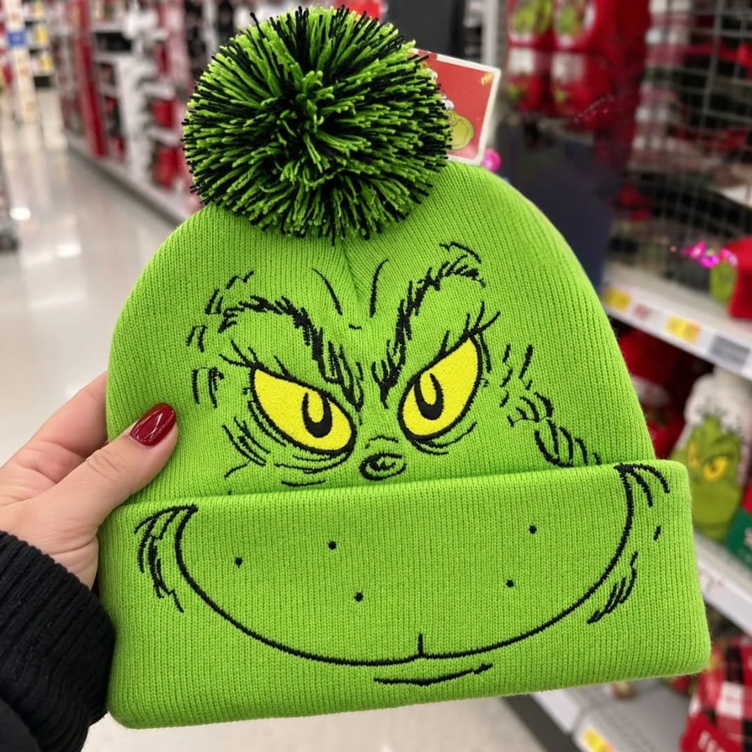 Hand holding a lime green Dr. Seuss Grinch beanie with embroidered face details and a green black pom-pom in a retail store