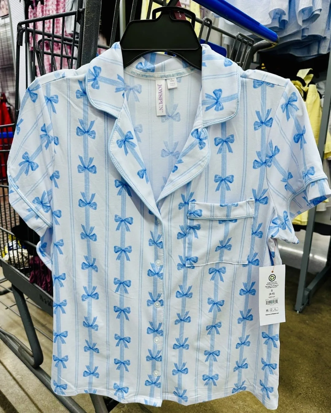 Joyspun short-sleeved pajama top with light blue bow and stripe pattern, hanging on a black hanger in a retail store, featuring a notched collar, chest pocket, and sustainable material tag.