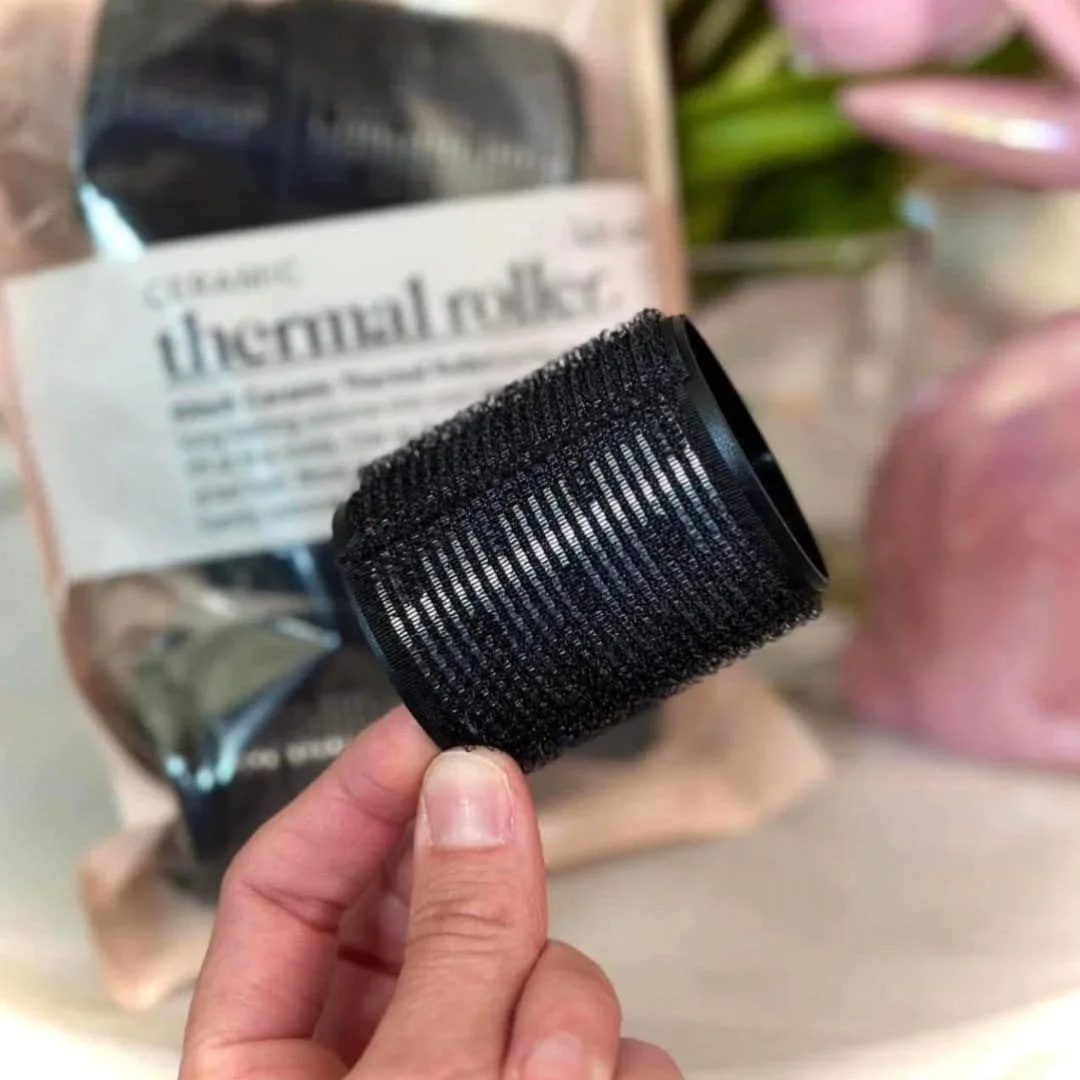 Kitsch ceramic thermal hair roller held by a hand, featuring a textured surface for long-lasting volume and smooth, shiny finish, with blurred packaging in the background.