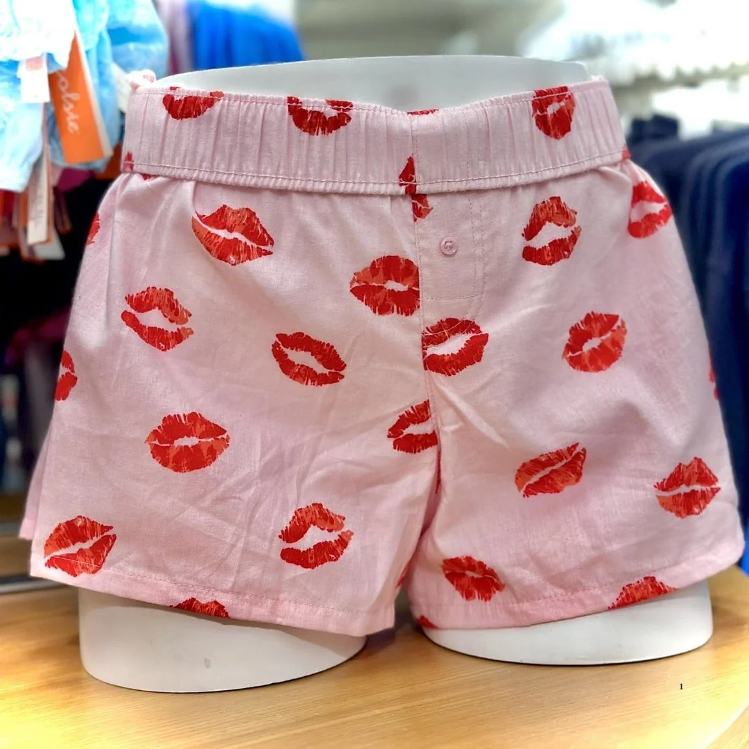 Light pink boxer shorts with a playful red lipstick kiss mark pattern, displayed on a white mannequin in a retail setting.