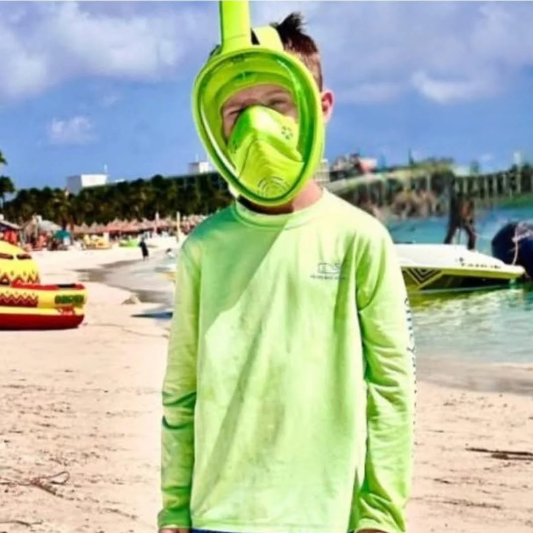 Young boy on a sunny beach wearing a lime green long-sleeve shirt and matching full-face snorkel mask, with a vibrant resort backdrop featuring palm trees, water toys, and turquoise ocean.