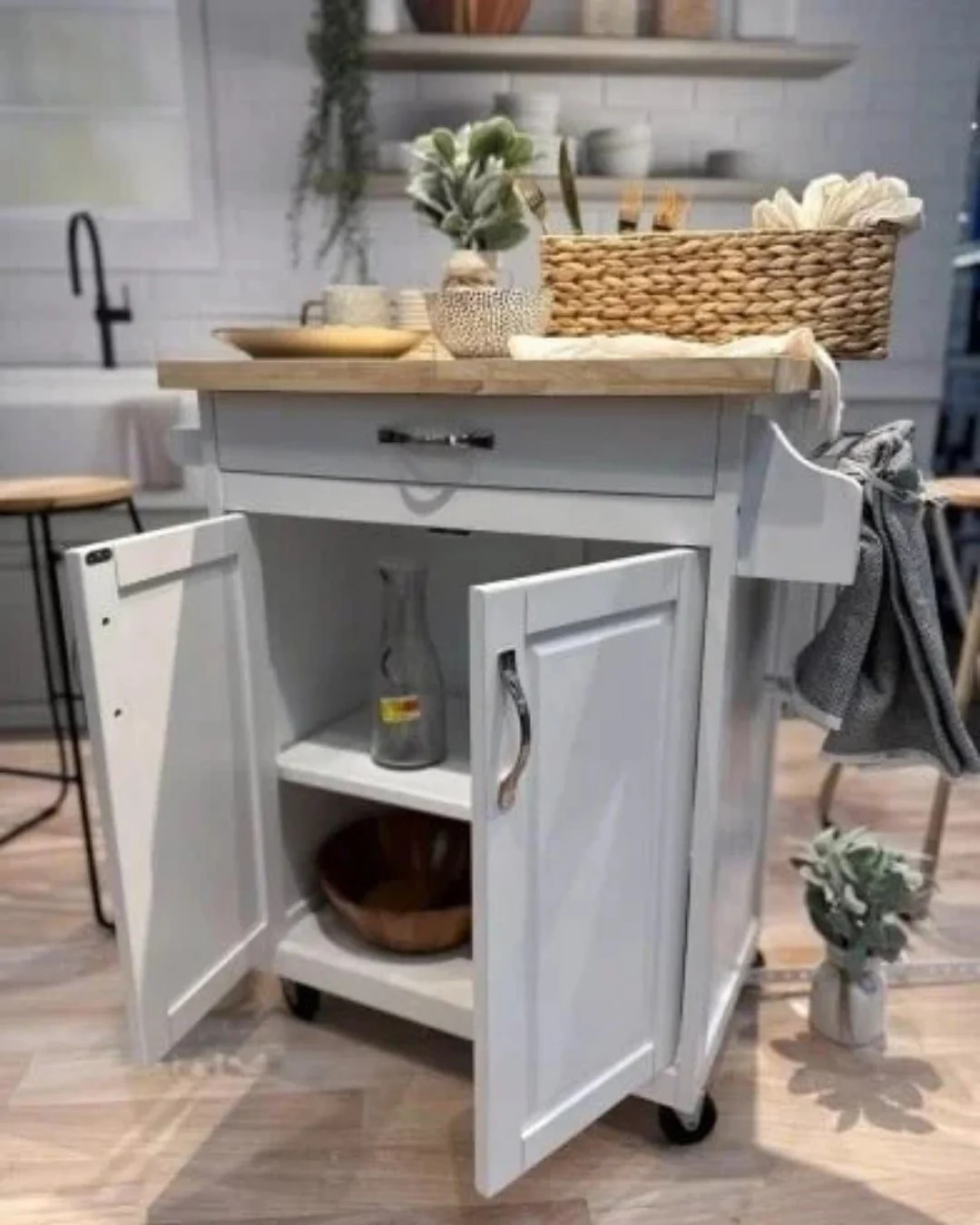 Mobile kitchen island with a light wood countertop, gray base, and open storage, styled with kitchen utensils, plants, and decor, set in a bright modern kitchen with a farmhouse sink and floating shelves.