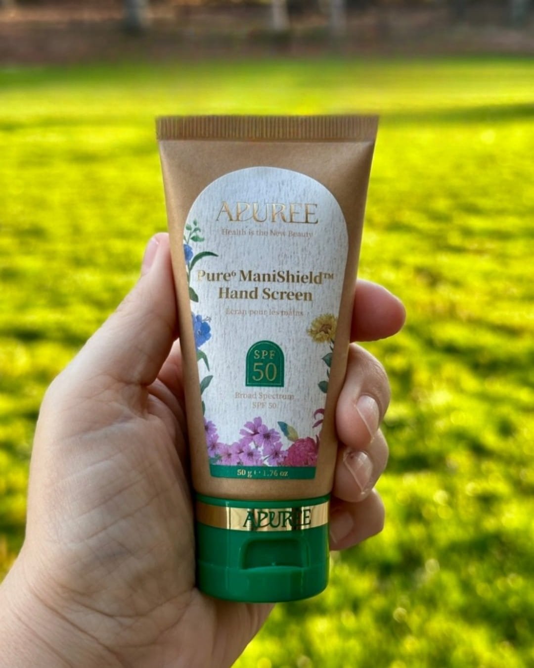 APUREE Pure ManiShield Hand Screen with SPF 50, eco-friendly kraft paper packaging, and floral illustrations, held outdoors against a blurred green lawn background.