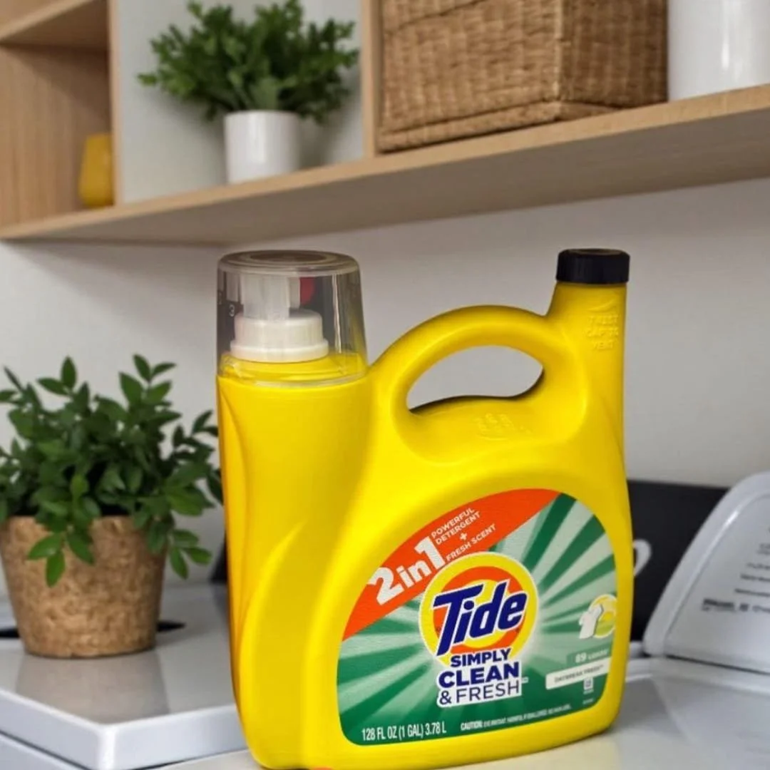 Tide Simply Clean & Fresh laundry detergent in a 128 fl oz yellow bottle with 89 loads capacity, displayed on a white surface in a clean laundry room with shelves, plants, and a woven basket in the background.