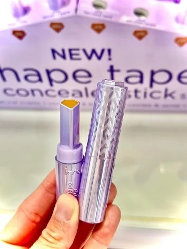 Close-up of a hand holding Tarte's new Shape Tape Concealer Stick, featuring an open hexagonal yellow-orange concealer and a textured purple and silver cap, against a blurred branded background.