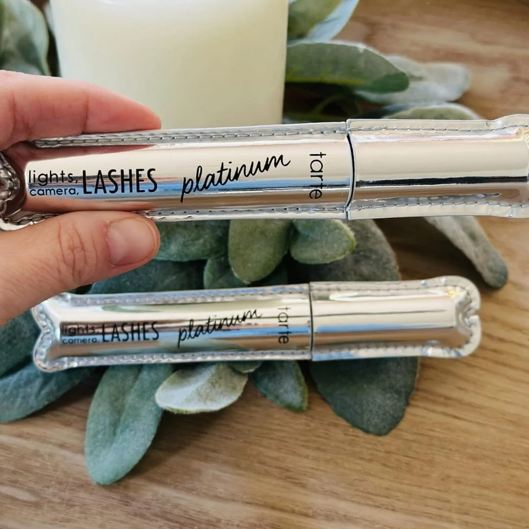Tarte Lights, Camera, Lashes Platinum Mascara with metallic silver packaging, showcased on a wooden surface with soft greenery and candle accents in the background.