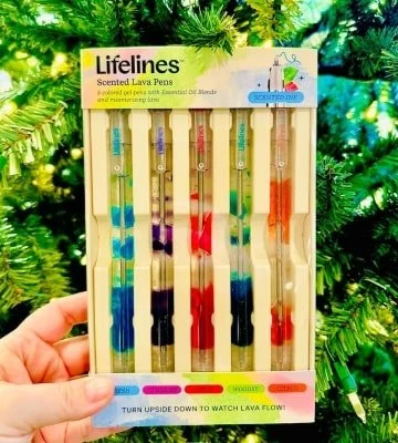 Box of Lifelines Scented Lava Pens featuring five colorful gel pens with essential oil blends and flowing lava, held against a festive green background with blurred lights.