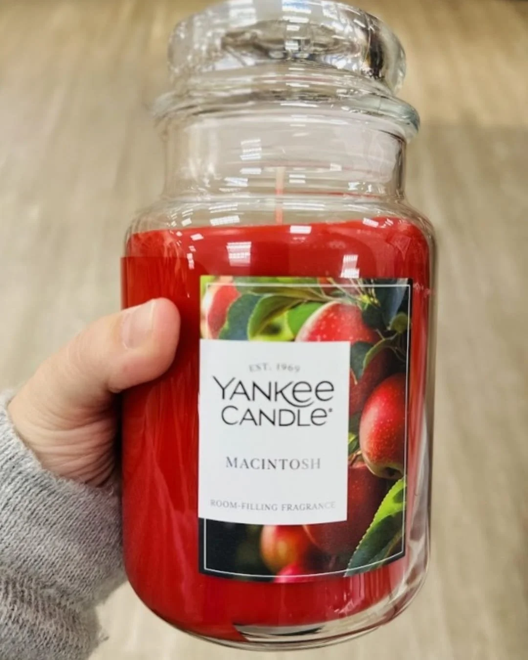 Yankee Candle Macintosh scent in a large glass jar with vibrant red wax, featuring a label with fresh red apples and green leaves, held by a hand wearing a grey knitted sweater.